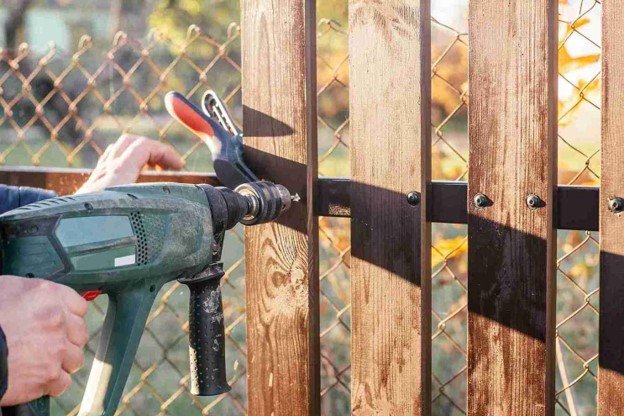 Contractor using a power drill to secure vertical wood boards during fence construction.